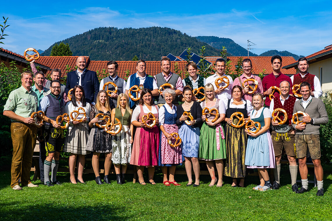 Gruppenfoto einiger Mitarbeiter in bayerischer Tracht und mit großer Breze im Garten der Firmenzentrale der itelio GmbH in Kiefersfelden mit Blick auf den Nußlberg.
